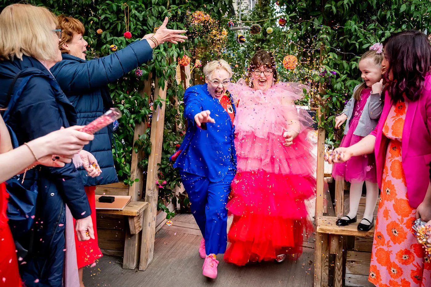 A very happy LGBTQ+ couple run through confetti. One is wearing a bright pink dress, and the other is wearing a bright blue suit.
