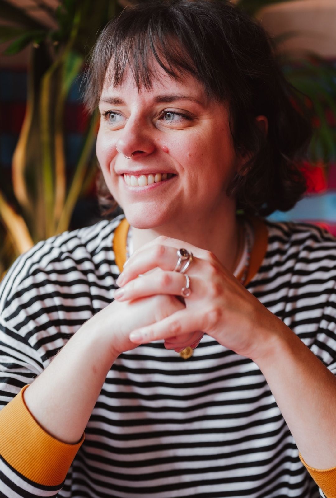 Hannah, an alternative-looking woman in her mid-30s smiles and looks into the distance. She has short brown hair, is wearing a black and white striped top with yellow trim, and silver rings on her fingers.