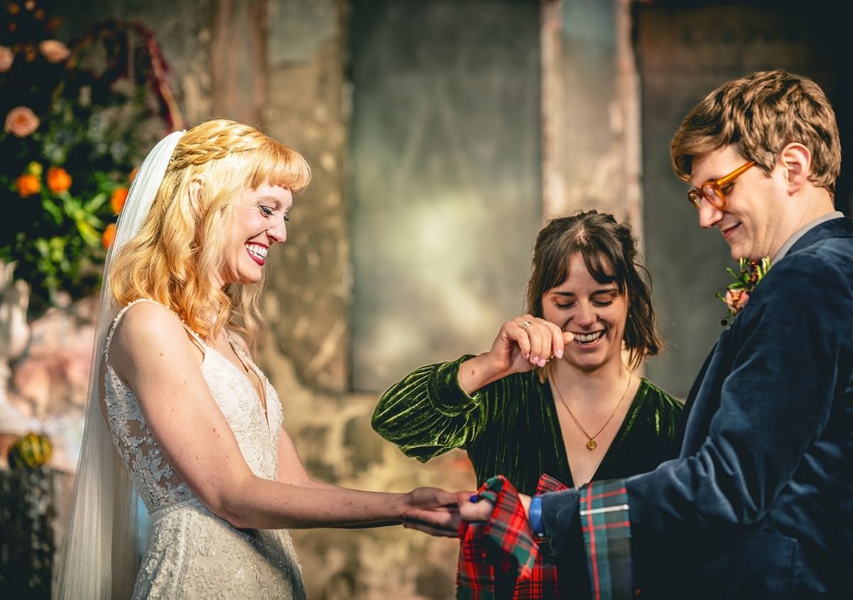 Hannah, an alternative wedding celebrant, is performing a handfasting with tartan ribbons. The couple are holding hands as Hannah ties the ribbon around them, and everyone is smiling and laughing.