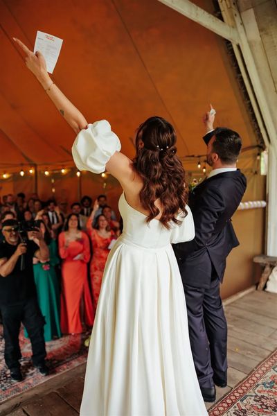 A happy couple have their hands in the air and are cheering as their guests look on and do the same. Their wedding is lead by Hannah, an alternative wedding celebrant.