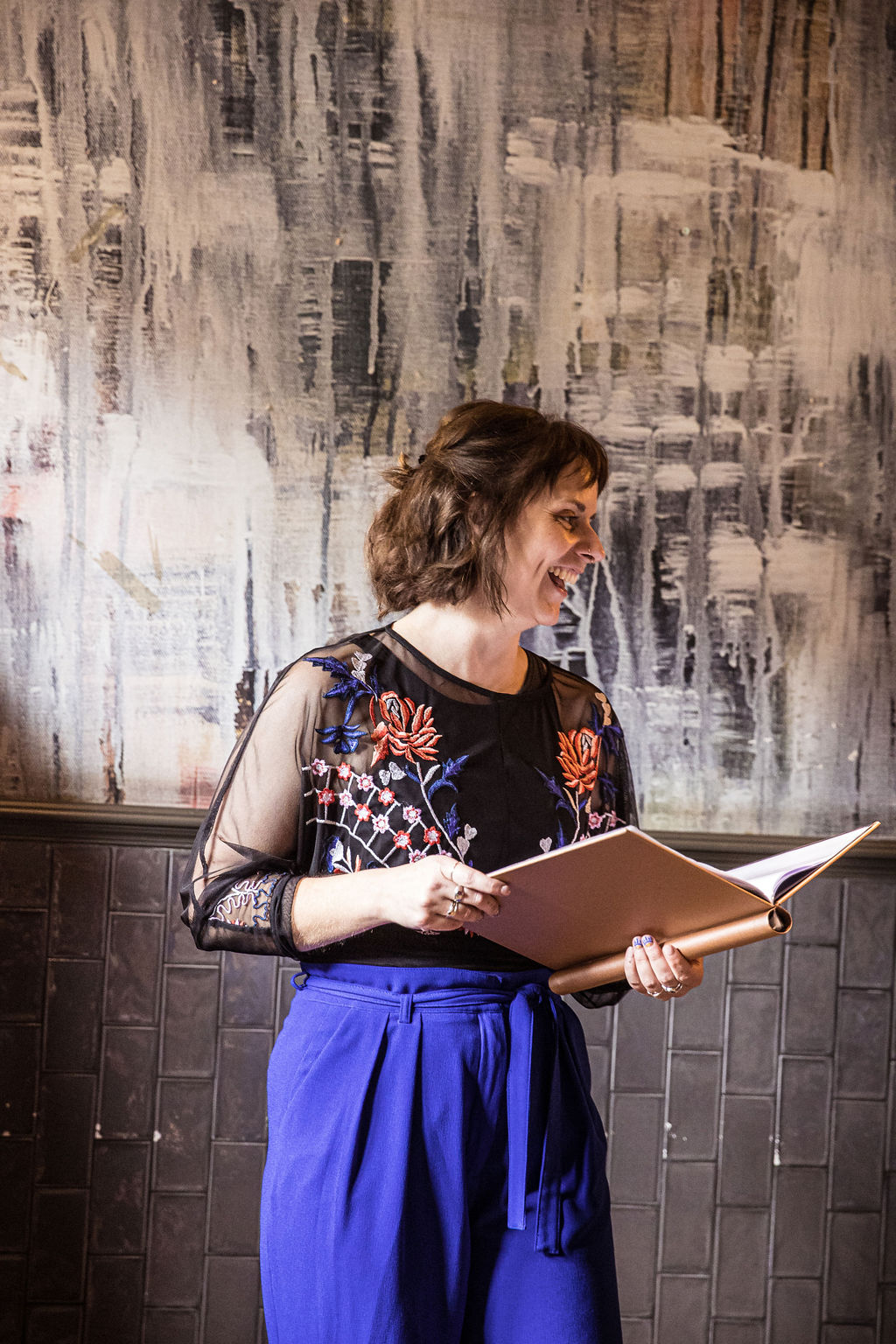 Hannah is reading aloud from her ceremony script. She is wearing bright blue trousers, a black top, and is smiling.