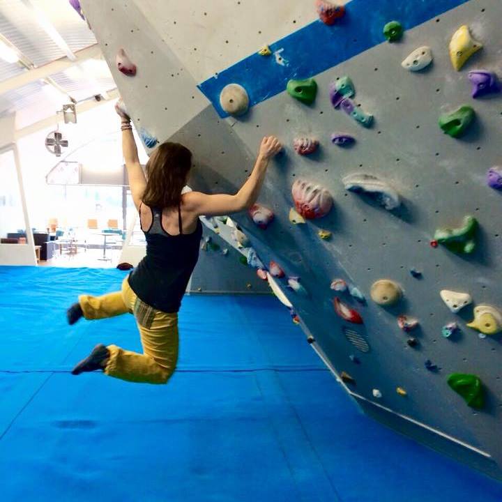 Hannah is holding onto some rock climbing holds at an indoor rock climbing centre whilst her body and legs swing in mid air.