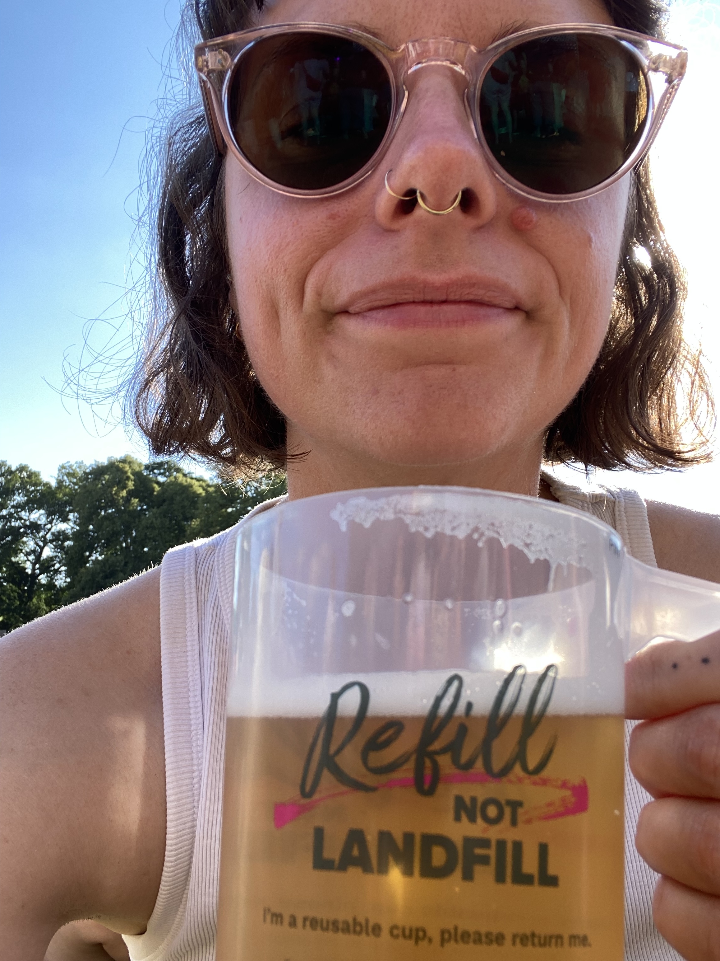 Hannah sitting in the sun, wearing sunglasses, drinking from a plastic pint glass. She looks very happy.