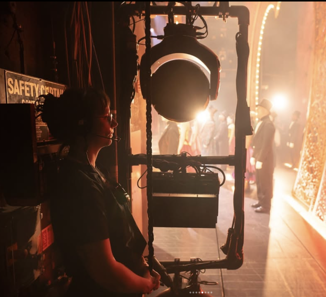 Hannah is standing backstage at the Piccadilly Theatre. She is dressed in black, wearing a headset, and watching the actors perform onstage. 